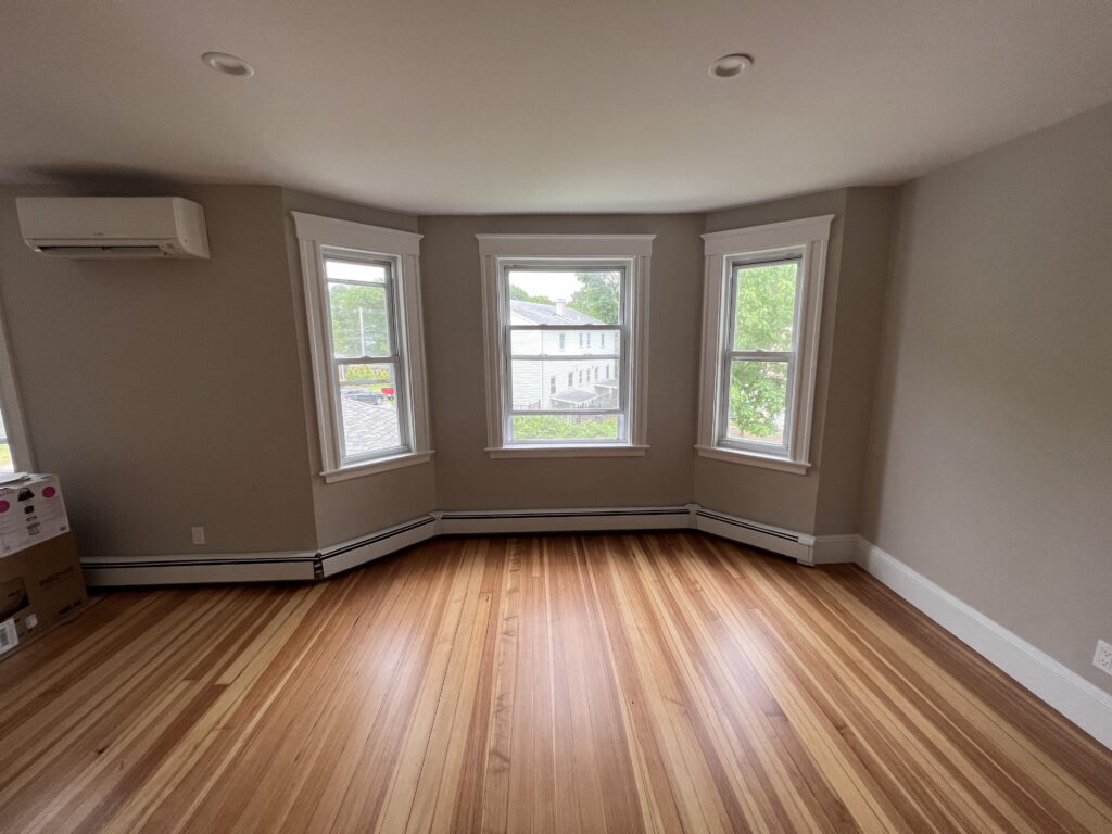 Bright room with three windows allowing natural light to accentuate the freshly refinished hardwood flooring, showcasing varying shades of wood. This space is part of a series detailing a hardwood floor refinishing project, with a focus on the floor's seamless finish and rich textures.