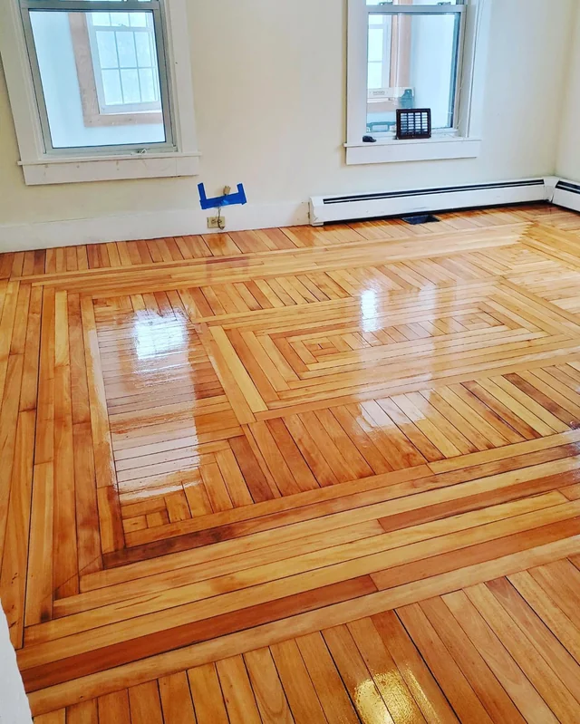 Intricate parquet hardwood flooring with a high-gloss finish, displaying expert craftsmanship in a sunlit room with minimalistic walls and two windows. This image forms part of a series highlighting the fifth project of hardwood floor refinishing, emphasizing the geometric design and the wood's natural golden tones.
