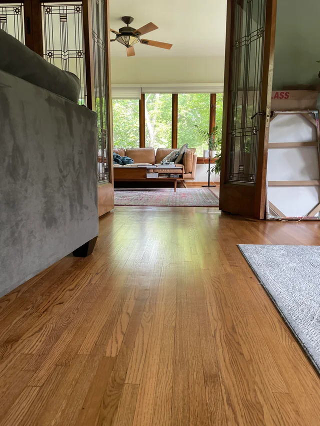 Warm and inviting room view with a focus on the lustrous oak hardwood flooring, capturing the ambiance of natural light and serene outdoor views in a home setting. This image is part of a series showcasing the meticulous process of hardwood floor refinishing, demonstrating the third project's attention to detail and craftsmanship.