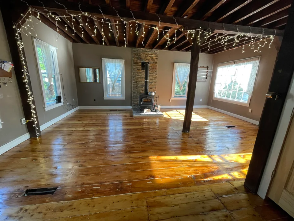 A spacious interior showcasing a recently polished wooden flooring with warm sunlight filtering through the windows, highlighting the room's rustic charm with exposed beams and a stone hearth. This Philadelphia home features rejuvenated hardwood floors as part of a detailed refinishing project.