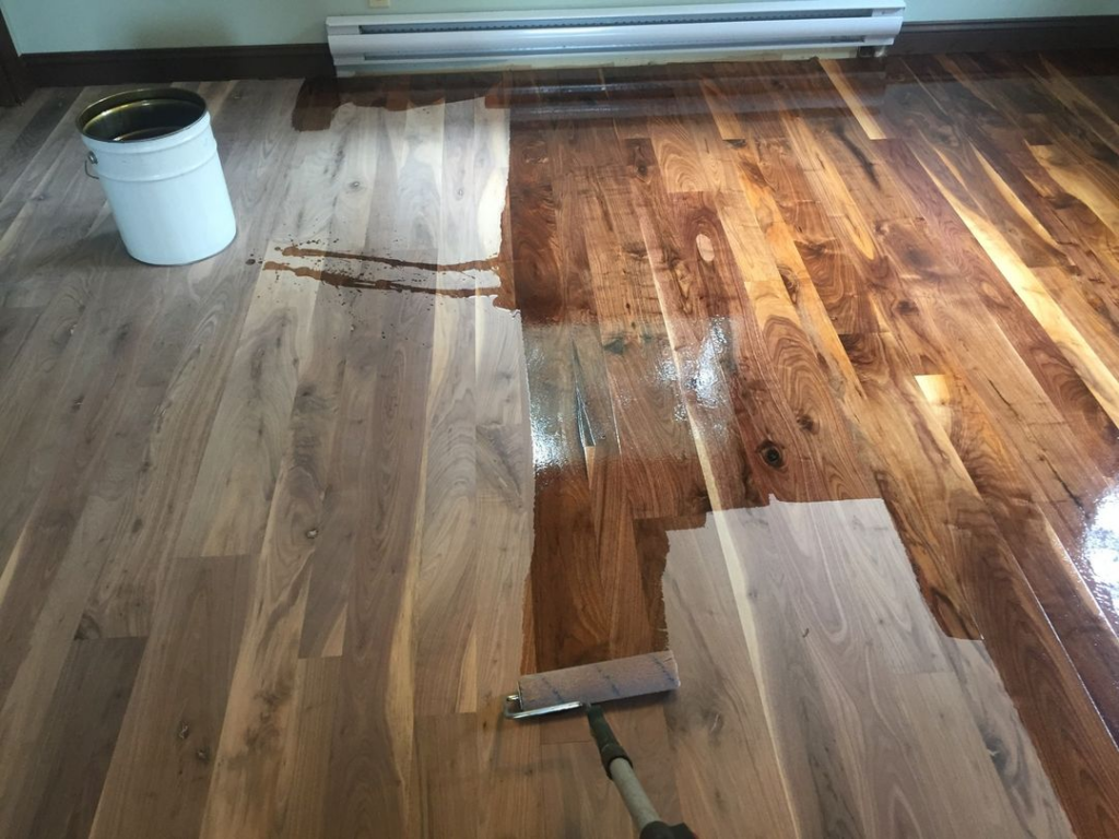 The process of refinishing a hardwood floor is underway in Philadelphia, with this image showing the eleventh project's application of a dark stain on the lighter wood, creating rich contrast and depth. A paintbrush, bucket, and partially treated floor indicate the transformation in progress.