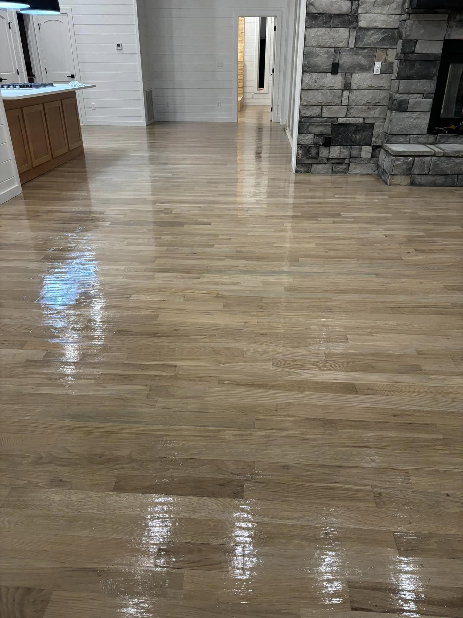 Modern kitchen with sleek, light-toned hardwood floors reflecting overhead lighting, part of a seventh project in a hardwood floor refinishing series. The floors extend towards a stone fireplace and white cabinetry, highlighting the contrast and brightness of the space.