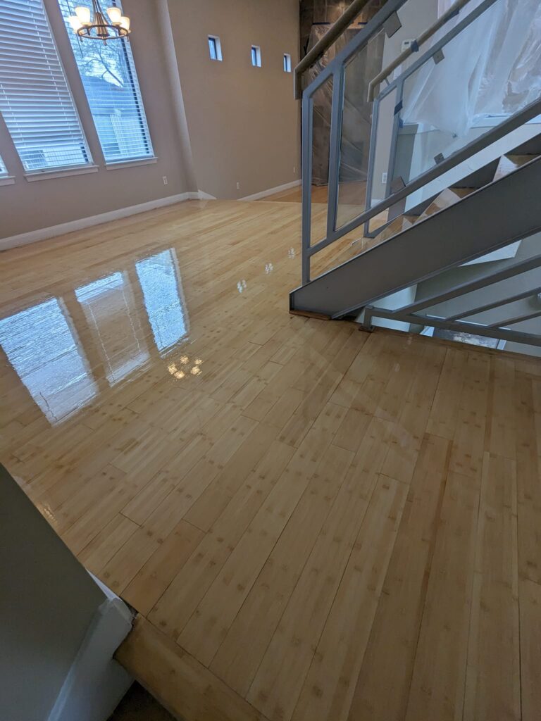 High-ceilinged room with a reflective, newly refinished hardwood floor, capturing the essence of Philadelphia craftsmanship in the eighth project. The light maple flooring contrasts with taupe walls and a contemporary staircase, enhancing the architectural details.